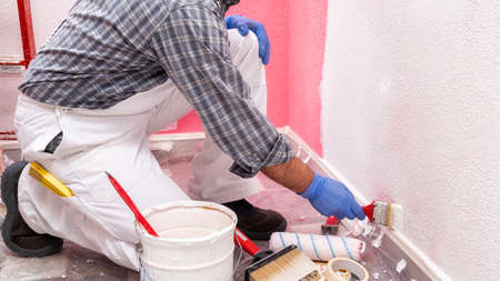 Caucasian house painter worker in white overalls, painting the pink wall with white paint using a small brush. Construction industry. Work safety.の写真素材