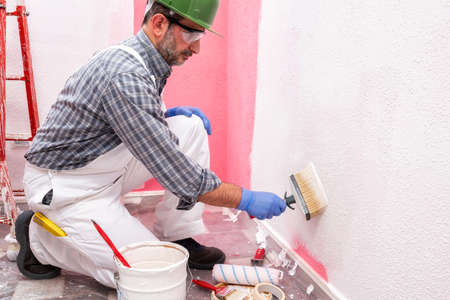 Caucasian house painter worker in white overalls, with helmet and goggles painting the pink wall with white paint using the brush. Construction industry. Work safety.の写真素材