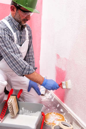 Caucasian house painter worker in white overalls, with helmet and goggles painting the pink wall with white paint using a small brush. Construction industry. Work safety.の写真素材