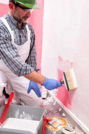 Caucasian house painter worker in white overalls, with helmet and goggles painting the pink wall with white paint using the brush. Construction industry. Work safety.の写真素材
