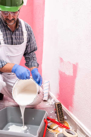 Caucasian house painter worker in white overalls, with helmet and goggles pour the white paint into the container. Construction industry. Work safety.の写真素材