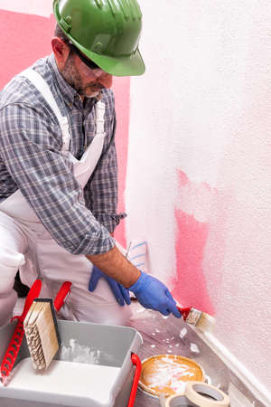 Caucasian house painter worker in white overalls, with helmet and goggles painting the pink wall with white paint using a small brush. Construction industry. Work safety.の写真素材