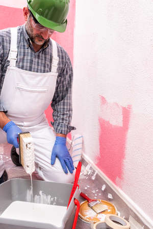 Caucasian house painter worker in white overalls, with helmet and goggles he prepares the white paint to paint the pink wall. Construction industry. Work safety.の写真素材