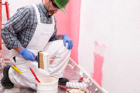 Caucasian house painter worker in white overalls, with helmet and goggles he prepares the white paint to paint the pink wall. Construction industry. Work safety.の写真素材