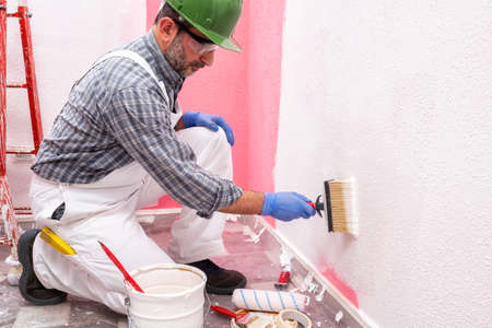 Caucasian house painter worker in white overalls, with helmet and goggles painting the pink wall with white paint using the brush. Construction industry. Work safety.の写真素材