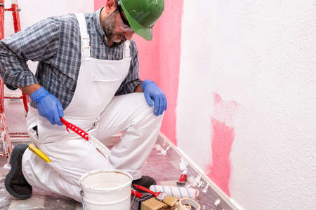 Caucasian house painter worker in white overalls, with helmet and goggles he prepares the white paint to paint the pink wall. Construction industry. Work safety.の写真素材