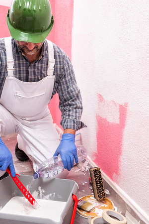 Caucasian house painter worker in white overalls, with helmet and goggles he prepares the white paint to paint the pink wall. Construction industry. Work safety.の写真素材