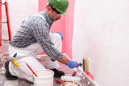 Caucasian house painter worker in white overalls, with helmet and goggles painting the pink wall with white paint using the brush. Construction industry. Work safety.の写真素材