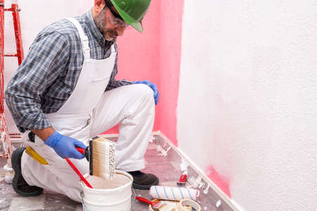 Caucasian house painter worker in white overalls, with helmet and goggles painting the pink wall with white paint using the brush. Construction industry. Work safety.の写真素材