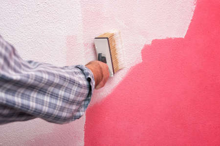 Caucasian house painter worker, painting the pink wall with white paint using the brush. Construction industry. Work safety.の写真素材