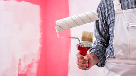 Caucasian house painter worker in white overalls, holding brush and roller for painting. Construction industry. Work tools.の写真素材