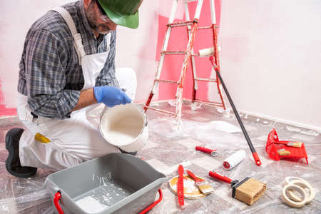 Caucasian house painter worker in white overalls, with helmet and goggles pour the white paint into the container. Construction industry. Work safety.の写真素材