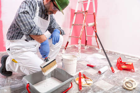 Caucasian house painter worker in white overalls, with helmet and goggles he prepares the white paint to paint the pink wall. Construction industry. Work safety.の写真素材