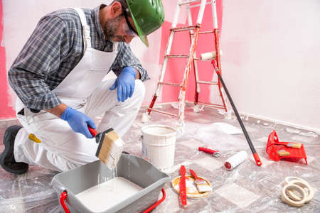 Caucasian house painter worker in white overalls, with helmet and goggles he prepares the white paint to paint the pink wall. Construction industry. Work safety.の写真素材