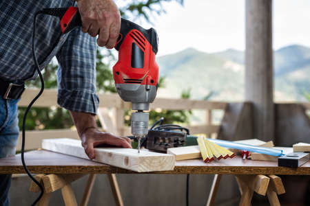 Adult craftsman carpenter with electric drill works makes a hole on a wooden table. Housework do it yourself. Stock photography.の写真素材