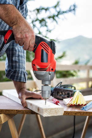 Adult craftsman carpenter with electric drill works makes a hole on a wooden table. Housework do it yourself. Stock photography.の写真素材