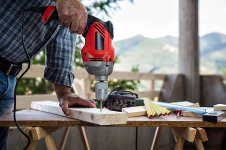 Adult craftsman carpenter with electric drill works makes a hole on a wooden table. Housework do it yourself. Stock photography.の写真素材