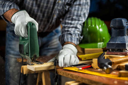 Adult carpenter craftsman wears protective leather gloves, with electric saw working on cutting a wooden table. Construction industry, housework do it yourself. Stock photography.の写真素材