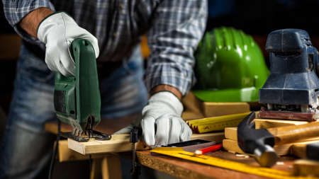 Adult carpenter craftsman wears protective leather gloves, with electric saw working on cutting a wooden table. Construction industry, housework do it yourself. Stock photography.の写真素材