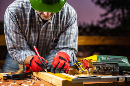 Adult carpenter craftsman wearing helmet and protective gloves, with a pencil and the carpenter's square trace the cutting line on a wooden table. Construction industry, housework do it yourself. Stock photography.の写真素材