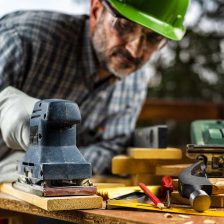 Adult carpenter craftsman wears protective leather gloves, with the electric sander smoothes a wooden table. Construction industry, housework do it yourself. Safety at work.の写真素材