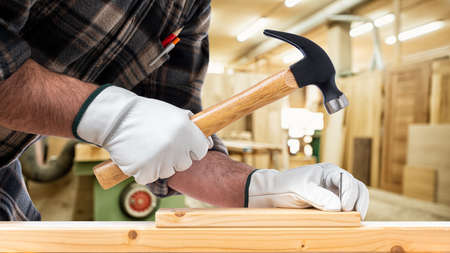 Close-up. Carpenter with his hands protected by gloves with a hammer and nails fixes a wooden board. Construction industry, carpentry workshop.の写真素材