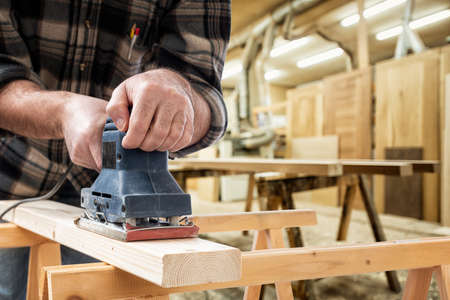 Close-up. Carpenter with the electric sander smoothes a wooden board. Construction industry, carpentry workshop.の写真素材