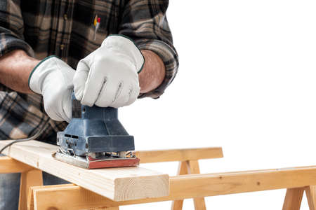 Close-up. Carpenter with his hands protected by gloves with the electric sander smoothes a wooden board. Construction industry. White background.の写真素材