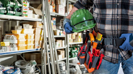 Electrician in the electrical component store wears protective gloves and holds helmet and goggles in hand. Construction industry, electrical system.の写真素材