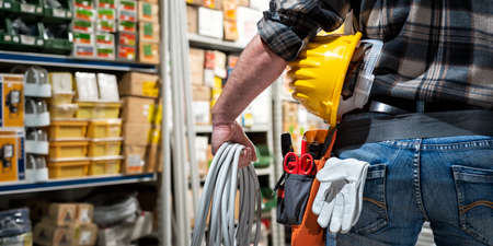 Electrician in the electrical component store holds the roll of electric cable in his hand, helmet with protective goggles. Construction industry, electrical system. View from behind.の写真素材