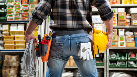 Electrician in the electrical component store holds the roll of electric cable in his hand, helmet with protective goggles. Construction industry, electrical system. View from behind.の写真素材