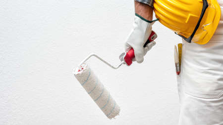 Caucasian house painter worker in white work overalls and with protective gloves, he holds the roller in his hand. Helmet and protective goggles. Work safety.の写真素材