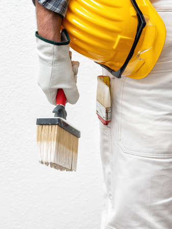 Caucasian house painter worker in white work overalls and with protective gloves, he holds the brush in his hand. Helmet and protective goggles. Work safety.の写真素材