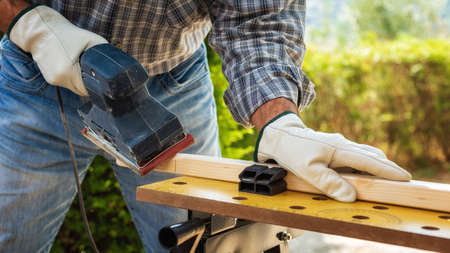Close-up. Carpenter with his hands protected by gloves with the electric sander smoothes a wooden board. Construction industry. Work safety.の写真素材
