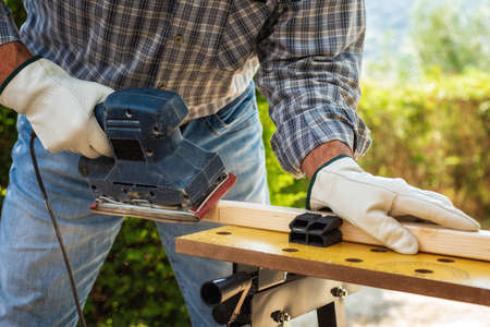 Close-up. Carpenter with his hands protected by gloves with the electric sander smoothes a wooden board. Construction industry. Work safety.の写真素材