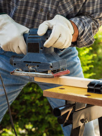 Close-up. Carpenter with his hands protected by gloves with the electric sander smoothes a wooden board. Construction industry. Work safety.の写真素材