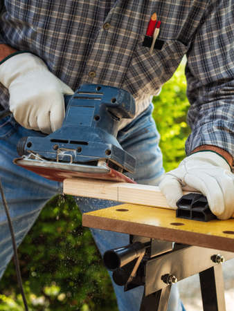 Close-up. Carpenter with his hands protected by gloves with the electric sander smoothes a wooden board. Construction industry. Work safety.の写真素材