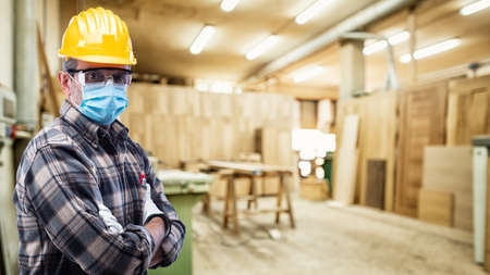 Carpenter worker in the carpentry workshop, wears helmet, goggles, leather gloves and surgical mask...