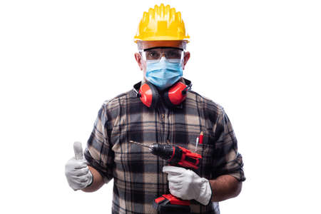 Carpenter worker isolated on white background wears surgical mask to prevent Coronavirus spread, makes OK sign with thumb up. Preventing Pandemic Covid-19 at the workplace.の写真素材