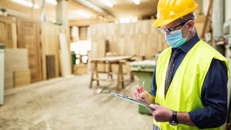 Engineer in carpentry workshop wears surgical mask to prevent Coronavirus spread, holds pen and notebook in hand. Preventing Pandemic Covid-19 at the workplace.の写真素材