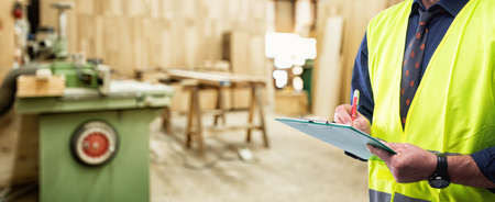 Engineer in carpentry workshop wears surgical mask to prevent Coronavirus spread, holds pen and notebook in hand. Preventing Pandemic Covid-19 at the workplace.の写真素材