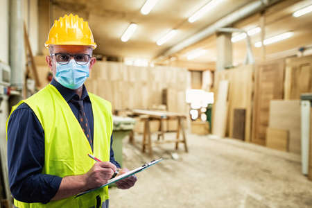 Engineer in carpentry workshop wears surgical mask to prevent Coronavirus spread, holds pen and notebook in hand. Preventing Pandemic Covid-19 at the workplace.の写真素材