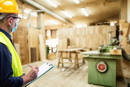 Engineer in carpentry workshop wears surgical mask to prevent Coronavirus spread, holds pen and notebook in hand. Preventing Pandemic Covid-19 at the workplace.の写真素材