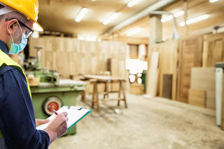 Engineer in carpentry workshop wears surgical mask to prevent Coronavirus spread, holds pen and notebook in hand. Preventing Pandemic Covid-19 at the workplace.の写真素材
