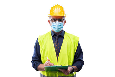 Engineer isolated on white background wears surgical mask to prevent Coronavirus spread, holds pen and notebook in hand. Preventing Pandemic Covid-19 at the workplace.の写真素材
