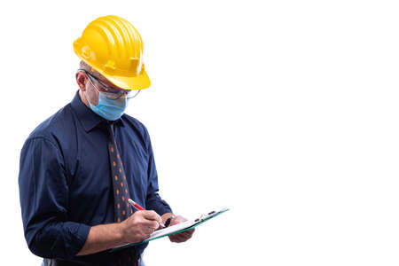 Engineer isolated on white background wears surgical mask to prevent Coronavirus spread, holds pen and notebook in hand. Preventing Pandemic Covid-19 at the workplace.の写真素材