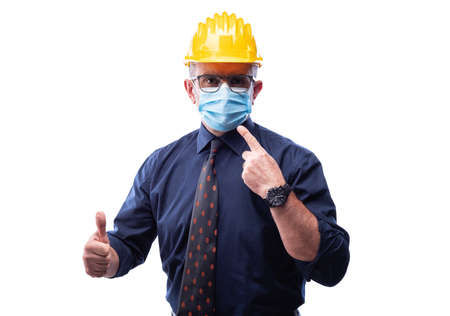 Engineer isolated on white background wears surgical mask to prevent Coronavirus spread, makes OK sign with thumb up. Preventing Pandemic Covid-19 at the workplace.の写真素材