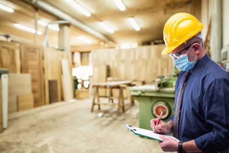 Engineer in carpentry workshop wears surgical mask to prevent Coronavirus spread, holds pen and notebook in hand. Preventing Pandemic Covid-19 at the workplace.の写真素材