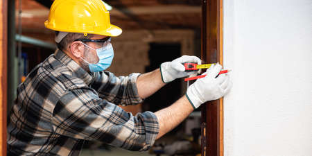 Carpenter worker at work repairs and installs a room door, wear the surgical mask to prevent Coronavirus infection. Preventing Pandemic Covid-19 at the workplace. Carpentry.の写真素材