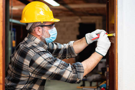 Carpenter worker at work repairs and installs a room door, wear the surgical mask to prevent Coronavirus infection. Preventing Pandemic Covid-19 at the workplace. Carpentry.の写真素材